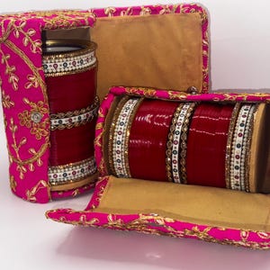 May include: Two pink velvet boxes with gold embroidery, each containing a set of red and white bangles. The boxes are open, revealing the bangles inside.