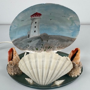 Peut inclure: Une lampe en coquillage avec une peinture de paysage marin représentant un phare sur une côte rocheuse. La lampe est faite de coquillages naturels et possède un cordon pour la brancher.