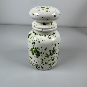 Op de afbeelding: Een witte keramische pot met een afgeronde deksel, versierd met onregelmatige groene verfspatten. De pot heeft een cilindrisch lichaam en een bolvormige deksel, wat een decoratieve en artistieke uitstraling creëert. Een uniek decoratiestuk.