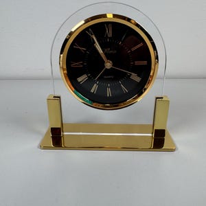 May include: A gold-toned quartz desk clock with a black face and gold Roman numerals. The clock has a clear, arched acrylic frame and a rectangular base. The clock face reads "Marco" and "Quartz".