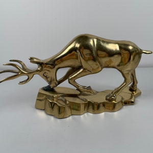 May include: A gold-toned brass sculpture of a deer with antlers, standing on a rocky base. The deer is depicted in a running pose, with its head lowered and its antlers pointing forward.