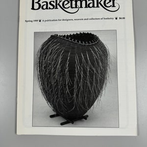 May include: A black and white magazine cover for Basketmaker, a publication for designers, weavers, and collectors of basketry. The cover features a black basket with white fringe, sitting on a stand.