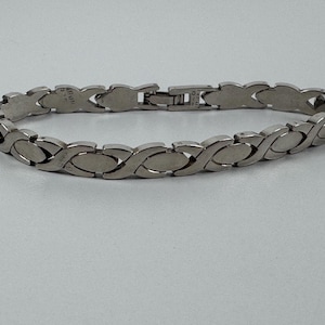 May include: A silver-toned bracelet with an interlocking oval design. The bracelet features a clasp closure and is displayed on a light gray surface. The jewelry piece is a stylish accessory.