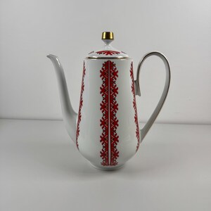 May include: White ceramic coffee pot with a red and black floral pattern. The pot has a curved handle, a long spout, and a gold-coloured finial on the lid. The design is symmetrical and runs vertically down the pot.