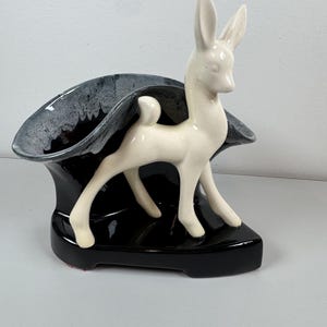May include: A vintage ceramic planter with a white deer figurine set against a black and grey glazed bowl. The deer stands on a glossy black base. The planter's design is a blend of mid-century modern and art deco styles, with a retro aesthetic.