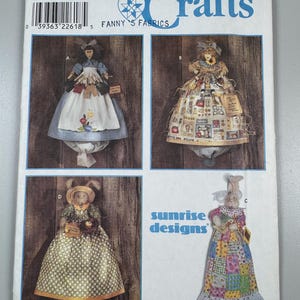 Op de afbeelding: Simplicity Crafts patroon met vier poppenontwerpen. De ontwerpen omvatten poppen met schorten, patchworkjurken en konijnenoren. Het patroon bevat de tekst "ONE SIZE" en "FANNY'S FABRICS".