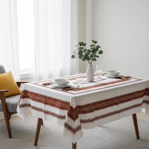 May include: A rectangular white tablecloth with a brown geometric border and trim. The table is set with white plates, bowls, and glasses. A white vase with greenery sits in the center. The table is on wooden legs.