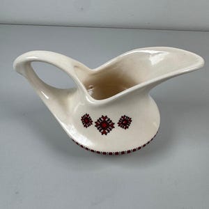 May include: A cream-coloured ceramic gravy boat featuring a wide pouring spout and a curved handle. The exterior is decorated with small, repeating geometric patterns in red and black, arranged in a horizontal band around the base and centre.