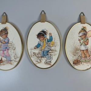 May include: Three oval-shaped wall decor pieces with gold frames. Each piece features a girl with a different activity: one is playing with ducks, one is playing tennis, and one is standing with a dog. The background is a light beige with brown accents.
