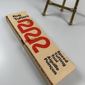 May include: A rectangular cardboard box for a Rug Tufters 222 Speed Tufting Tool Aiguille Poinçon. The packaging is tan with black text and a large red number 222 printed on the top surface. The box is positioned diagonally on a white surface.