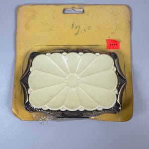 May include: A rectangular soap dish featuring a cream-coloured, petal-shaped centre insert surrounded by a dark, antiqued bronze-toned metal frame with decorative edges. The item is mounted on a yellow cardboard backing with a hole for hanging.