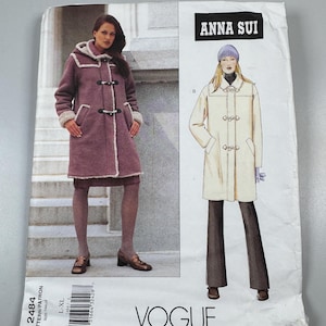 May include: A Vogue Patterns sewing pattern envelope for an Anna Sui coat, numbered 2484 in size L-XL. The front displays a photograph of a mauve hooded coat with toggle closures and a white illustration of a cream-colored coat with similar toggles.
