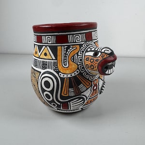 May include: A ceramic mug with a red rim and a black, white, orange, and yellow design. The mug features a stylised animal face with a wide open mouth.