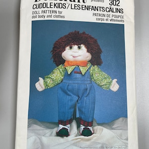 Puede incluir: Patrón de muñeca Dollcraft para un niño de peluche llamado "Skoooter/Coquin". El patrón es para un cuerpo de muñeca y ropa. La muñeca es un niño de pelo castaño con una camisa verde y blanca, un mono azul y zapatos rojos y negros. El número de patrón es 302.