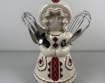 May include: A cream-colored ceramic figurine shaped like a person wearing a bonnet and dress, decorated with red embroidered-style patterns. The figurine features two metal wire whisks and a metal serving fork tucked into its arms as accessories.