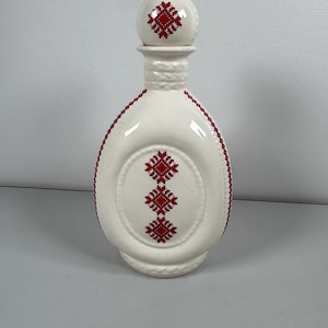 May include: A white ceramic bottle with a decorative red cross-stitch pattern. The bottle has a round stopper with a matching design. The body of the bottle features an oval design with three red cross-stitch patterns.
