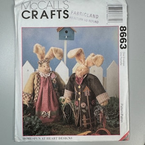 May include: A McCall's Crafts pattern for making two bunny dolls. The pattern is called "Homespun at Heart Designs" and the pattern number is 8663. The dolls are dressed in patchwork clothing and are standing in front of a white picket fence.