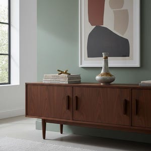 Può includere: Un mobile in legno in stile moderno di metà secolo con tre ante e un vaso sopra. Un'opera d'arte astratta incorniciata è appesa alla parete sopra il mobile. La stanza ha una palette di colori neutri con una parete verde e un tappeto grigio chiaro.