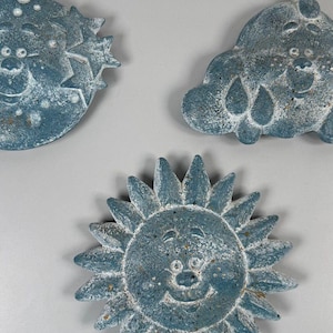 Puede incluir: Tres placas de pared texturizadas de color azul grisáceo con motivos celestiales. Una placa es un sol con rayos, otra es una luna con estrellas y la tercera es una nube con gotas de lluvia. Cada pieza tiene un acabado desgastado.