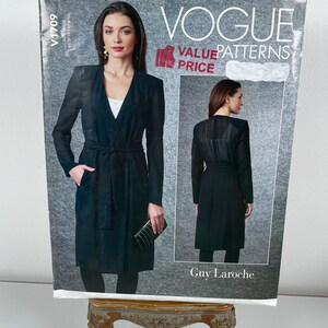 May include: A Vogue Patterns sewing pattern for a black wrap coat. The pattern packet shows a model wearing the coat, with the text "Vogue Value Patterns" and "Guy Laroche". The pattern is displayed on a decorative stand.