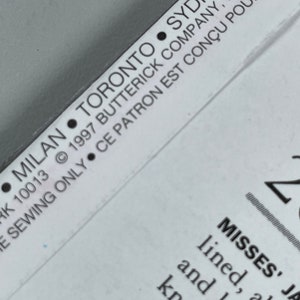 May include: A close-up of a white sewing pattern envelope with black text. The text reads: "&bull; MILAN &bull; TORONTO &bull; SYDNEY &bull; RK 10013 &copy; 1997 BUTTERICK COMPANY, INC. &bull;  E SEWING ONLY CE PATRON EST CON&Ccedil;U POUR L'USAG MISSES' JA lined, above and long knee or elbow abo f"