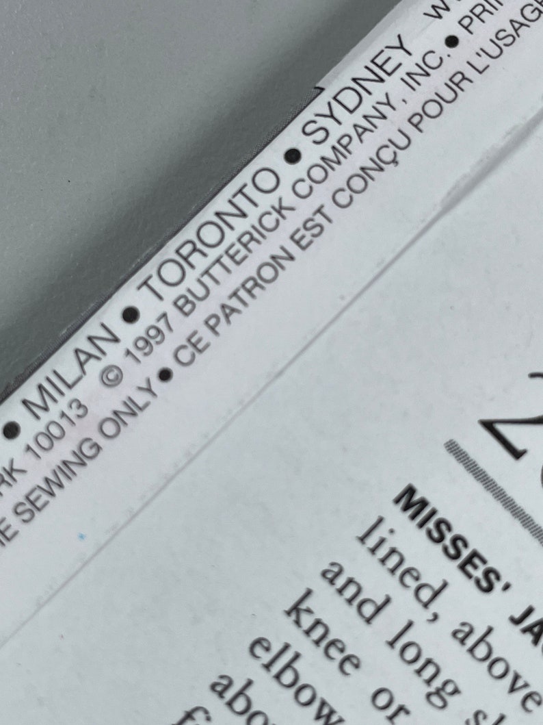 May include: A close-up of a white sewing pattern envelope with black text. The text reads: "&bull; MILAN &bull; TORONTO &bull; SYDNEY &bull; RK 10013 &copy; 1997 BUTTERICK COMPANY, INC. &bull;  E SEWING ONLY CE PATRON EST CON&Ccedil;U POUR L'USAG MISSES' JA lined, above and long knee or elbow abo f"