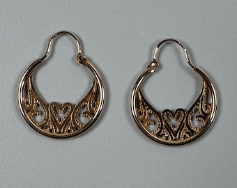 May include: A pair of gold-colored hoop earrings. Each earring features a heart-shaped filigree design at the bottom, with intricate details. The earrings have a classic hoop shape with a simple clasp.