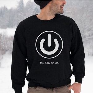 May include: Black crew neck sweatshirt featuring a white power button graphic and the text "You turn me on." The design is simple and bold. The sweatshirt is made of a soft material.