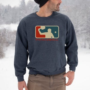 May include: A navy blue sweatshirt with a pickleball player graphic inside a rectangle. The word "PICKLEBALL" is printed in red below. The sweatshirt is worn by a person in a snowy outdoor setting, with a black hat.