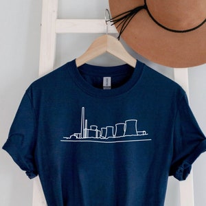 May include: A navy blue t-shirt with a white outline of a power plant skyline. The shirt is hanging on a white ladder with a brown hat on top.