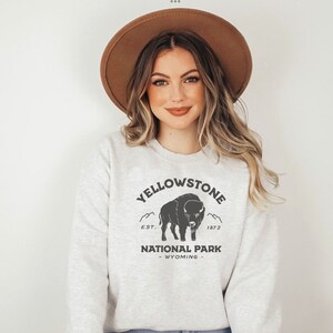May include: A light gray sweatshirt with the text "YELLOWSTONE NATIONAL PARK WYOMING" and a bison graphic. The sweatshirt has a crew neck and long sleeves. The text and graphic are in black.