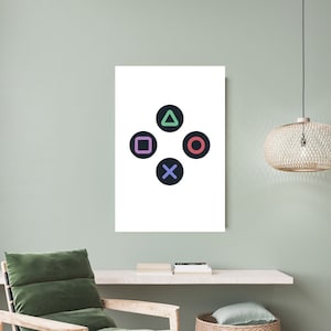 May include: A white canvas print featuring the iconic PlayStation controller button symbols: a green triangle, a purple square, a red circle, and a blue X. The symbols are set against a black background. The print is displayed in a room with a green chair.