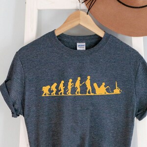 以下が含まれることがあります： 暗いヘザーグレーのTシャツに、黄色で描かれた人間の進化のグラフィックがプリントされています。進化の最終段階は、椅子に座ってビデオゲームをしている人です。