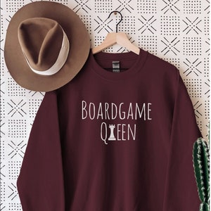 Può includere: Felpa color borgogna con testo bianco che recita "Boardgame Queen" e un'icona di regina degli scacchi bianca.