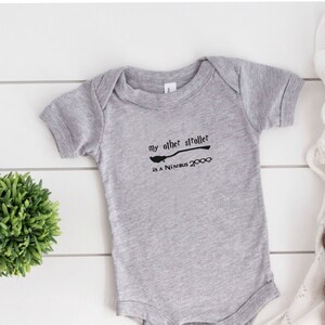 Può includere: Un body grigio per bambini con la scritta "my other stroller is a Nimbus 2000" in nero. Il body è accanto a una piccola pianta verde, una coperta bianca e un paio di scarpine marroni.