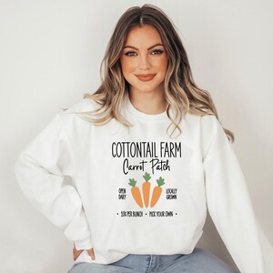 Puede incluir: Sudadera blanca con el texto "COTTONTAIL FARM Carrot Patch" en negro. El diseño incluye tres zanahorias naranjas con hojas verdes. El texto adicional dice "OPEN DAILY", "LOCALLY GROWN" y "10¢ PER BUNCH - PICK YOUR OWN".