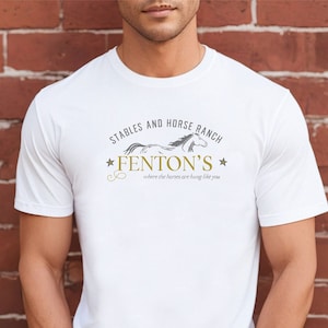 May include: White t-shirt with the text "Stables and Horse Ranch Fenton's where the horses are hung like you" in gray and gold. The shirt features a horse graphic. The model is wearing blue jeans.