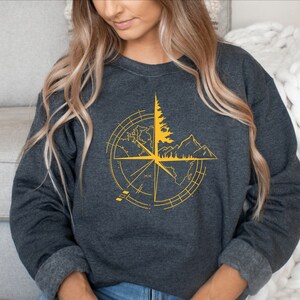 May include: Dark gray sweatshirt featuring a yellow compass design with a tree, mountains, and world map elements. The design is centered on the chest. The sweatshirt has long sleeves and a crew neck.