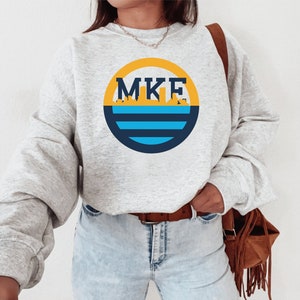Könnte beinhalten: Ein graues Sweatshirt mit einem gelben, blauen und schwarzen Kreis-Motiv mit dem Text "MKE" und einer Skyline-Silhouette.
