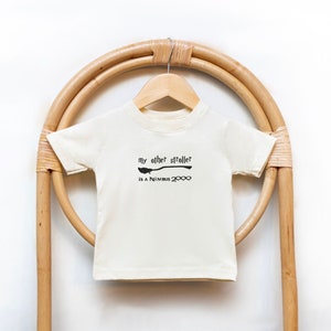 May include: Cream-colored short-sleeved t-shirt with the text "my other stroller is a Nimbus 2000" in black. The design includes a broom graphic. The shirt is hanging on a wooden hanger against a light brown, woven background.