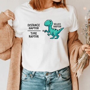 May include: White t-shirt with a teal cartoon velociraptor illustration. The shirt features the text "Distance Raptor / Time Raptor = Veloci Raptor" in black. The person is wearing blue jeans and a tan cardigan.
