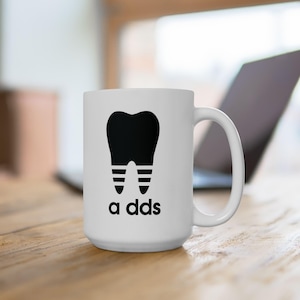 May include: White ceramic mug featuring a black tooth graphic and the text "a dds". The tooth design has striped roots, resembling a sports logo. The mug has a large handle and is likely for beverages.