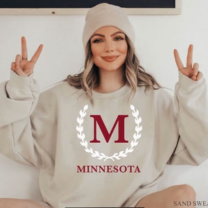 May include: Sand-colored sweatshirt with a maroon "M" inside a white laurel wreath, and the word "MINNESOTA" below. The sweatshirt is a neutral color and has long sleeves. The text "SAND SWEATSHIRT" is at the bottom right.