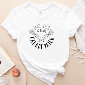 May include: White t-shirt with a black graphic featuring crossed carrots and the text "FARM FRESH 10 CENTS CARROT PATCH" in a circular design. The shirt is knotted at the bottom.