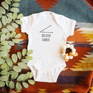 May include: White baby onesie with the text "acute baby" and a graphic of an acute angle. A wooden and white bead teether is next to the onesie. The onesie is on a colorful patterned blanket with green leaves.