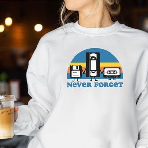 Puede incluir: Sudadera blanca con un diseño retro que presenta un disquete, una cinta VHS y una cinta de cassette tomados de la mano bajo un arco azul y amarillo. El texto "Never Forget" está impreso debajo en azul.