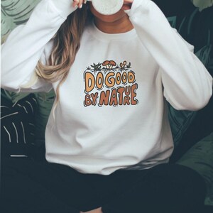 Puede incluir: Sudadera blanca con el texto "DO GOOD BY NATURE" en naranja y marrón, con un gráfico de setas y plantas. La sudadera es de cuello redondo y manga larga.