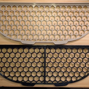 Puede incluir: Dos rejillas semicirculares con patrón de panal. Una es de color gris claro, la otra negra. Ambas están hechas de un material rígido y se colocan sobre una superficie de madera. Las rejillas tienen aberturas hexagonales.