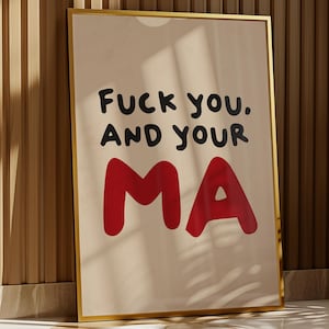 May include: A framed art print with the text "Fuck you. AND YOUR MA" in black and red lettering. The print has a beige background and a gold frame. The artwork is leaning against a wooden wall.