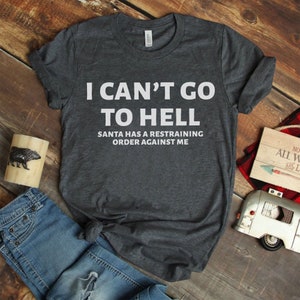 May include: A dark gray t-shirt with the text "I can't go to hell. Santa has a restraining order against me." printed in white.